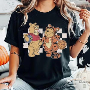 May include: Black t-shirt featuring a vintage-style graphic of two cartoon characters dressed in Halloween costumes. One character is in a bear costume, and the other is in a tiger costume, holding a pumpkin. The background has a checkered pattern.