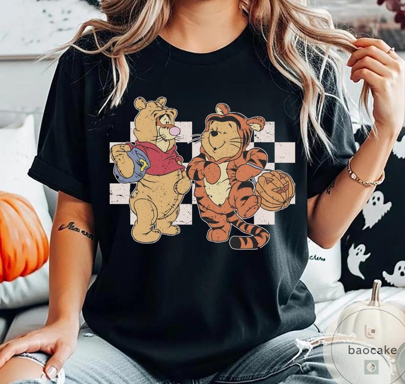 May include: Black t-shirt featuring a vintage-style graphic of two cartoon characters dressed in Halloween costumes. One character is in a bear costume, and the other is in a tiger costume, holding a pumpkin. The background has a checkered pattern.