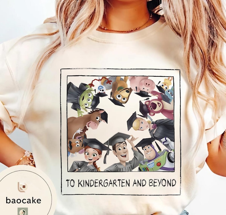 May include: Cream-coloured t-shirt with a framed graphic of Toy Story characters wearing graduation caps. The text "TO KINDERGARTEN AND BEYOND" is printed below the image. The shirt has a crew neck and short sleeves.