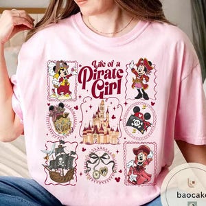 May include: Pink t-shirt featuring a design with the text "Life of a Pirate Girl" and illustrations of a pirate-themed Minnie Mouse. The design includes a pirate ship, treasure chest, and a castle.