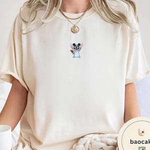 May include: A cream-colored short-sleeve t-shirt featuring a small, centered graphic of the blue character Stitch wearing black mouse ears and holding a brown cookie. The shirt has a crew neckline and is worn with a gold-toned chain necklace.