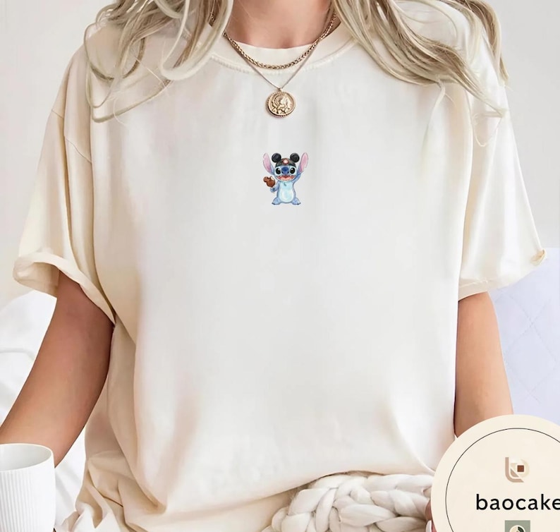 May include: A cream-colored short-sleeve t-shirt featuring a small, centered graphic of the blue character Stitch wearing black mouse ears and holding a brown cookie. The shirt has a crew neckline and is worn with a gold-toned chain necklace.