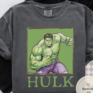 May include: Dark gray t-shirt featuring a green illustration of the Hulk in a fighting pose, set against a green background. The word "HULK" is printed in green below the image. The shirt has a crew neck and short sleeves.