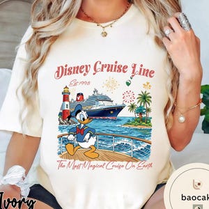 K&ouml;nnte beinhalten: Elfenbeinfarbenes T-Shirt mit einer Grafik von Donald Duck auf einem Kreuzfahrtschiff. Das Design umfasst einen Leuchtturm, Palmen und den Text "Disney Cruise Line" und "The Most Magical Cruise On Earth."