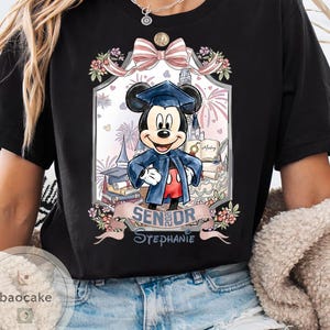 May include: Black t-shirt featuring a graduation-themed Mickey Mouse graphic. Mickey is wearing a graduation cap and gown, surrounded by books, fireworks, and a banner that reads "Senior Stephanie."