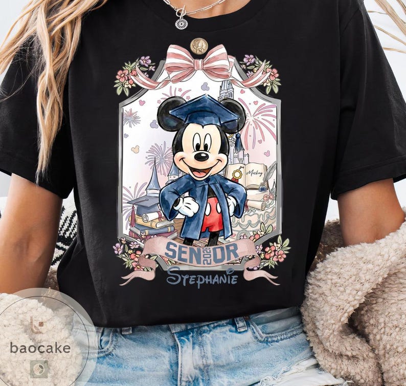 May include: Black t-shirt featuring a graduation-themed Mickey Mouse graphic. Mickey is wearing a graduation cap and gown, surrounded by books, fireworks, and a banner that reads "Senior Stephanie."