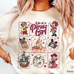 May include: Cream-colored t-shirt featuring a "Life of a Pirate Girl" design. The design includes illustrations of Minnie Mouse in pirate attire, a treasure chest, a pirate ship, a castle, and other pirate-themed elements.