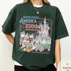 May include: Dark green t-shirt featuring a graphic celebrating the 250th anniversary of the United States of America. The design includes a castle, fireworks, and cartoon characters like Mickey Mouse and Donald Duck.