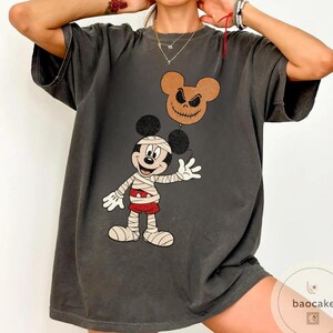 May include: Dark gray t-shirt featuring a cartoon mummy character holding a balloon with a jack-o'-lantern face. The mummy is wearing bandages and red shorts. The balloon is brown with a stitched mouth and eyes. The shirt has a vintage, distressed look.
