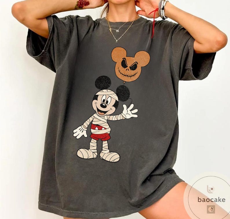 May include: Dark gray t-shirt featuring a cartoon mummy character holding a balloon with a jack-o'-lantern face. The mummy is wearing bandages and red shorts. The balloon is brown with a stitched mouth and eyes. The shirt has a vintage, distressed look.