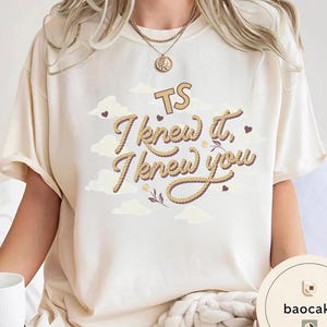 K&ouml;nnte beinhalten: Ein cremefarbenes Kurzarm-T-Shirt mit einem Grafikdesign, das den Text "TS I knew it, I knew you" in einer seilartigen Schriftart zeigt. Das Design enth&auml;lt kleine braune Herzen, helle Wolken und florale Akzente.