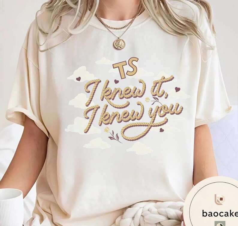 K&ouml;nnte beinhalten: Ein cremefarbenes Kurzarm-T-Shirt mit einem Grafikdesign, das den Text "TS I knew it, I knew you" in einer seilartigen Schriftart zeigt. Das Design enth&auml;lt kleine braune Herzen, helle Wolken und florale Akzente.