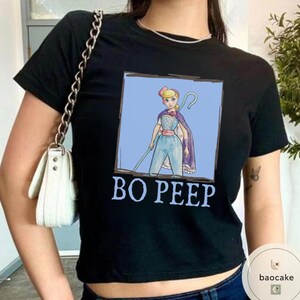K&ouml;nnte beinhalten: Ein schwarzes Kurzarm-T-Shirt mit einem quadratischen Aufdruck einer Zeichentrickfigur mit blonden Haaren, die ein blaues Outfit und einen violetten Umhang tr&auml;gt und einen Hirtenstab h&auml;lt. Der Text BO PEEP ist in hellblauen Buchstaben darunter gedruckt.