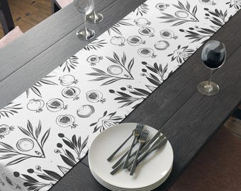 May include: A black and white table runner with a botanical design featuring pomegranates and leaves. The runner is placed on a dark wooden table set for a meal, with plates, silverware, and wine glasses. A bowl of fruit sits at the center.