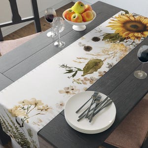 Op de afbeelding: Een eettafel met een witte bloemen tafelloper met een grote zonnebloem en andere botanische elementen. De tafel is gedekt met witte borden, bestek, wijnglazen en een kom met appels. De tafel en stoelen zijn van donker hout.
