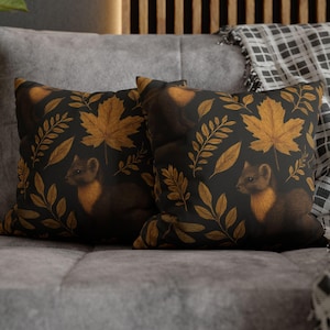 Puede incluir: Dos cojines cuadrados con un fondo negro que presenta un diseño de comadreja dorada y hojas de otoño. Los cojines están colocados sobre un sofá gris con una manta a cuadros en el fondo.