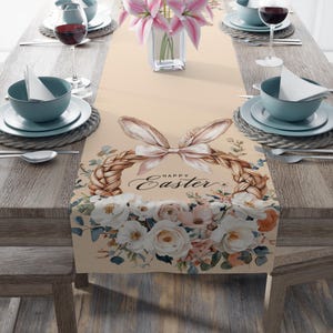 Op de afbeelding: Een beige Paas tafelloper met een bloemen- en konijnenoordesign. De loper heeft een gevlochten mand met konijnenoren en de woorden "Happy Easter". Witte en roze bloemen bevinden zich aan de onderkant.