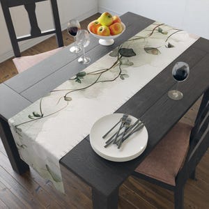 Puede incluir: Una mesa de comedor de madera oscura con un camino de mesa floral, platos blancos, tenedores, copas de vino y un cuenco de manzanas. El camino de mesa presenta un diseño de acuarela con hojas y vides verdes.