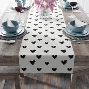 May include: A cream-colored table runner with a repeating pattern of black hearts. The runner is placed on a wooden table set for a meal, with plates, bowls, silverware, and wine glasses. A vase of pink flowers is in the center.