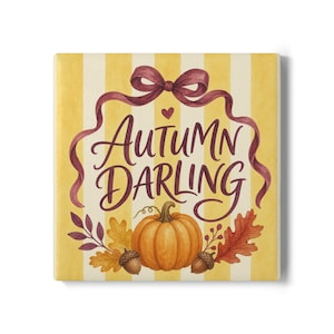 May include: A square decorative tile featuring a yellow and white vertical striped background. The text "AUTUMN DARLING" is written in dark purple script, framed by a purple ribbon bow, an orange pumpkin, two brown acorns, and fall leaves.