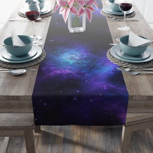 May include: A table runner featuring a vibrant nebula design in shades of blue, purple, and black. The runner is placed on a wooden table set with plates, silverware, and wine glasses. A vase of pink flowers sits in the center.