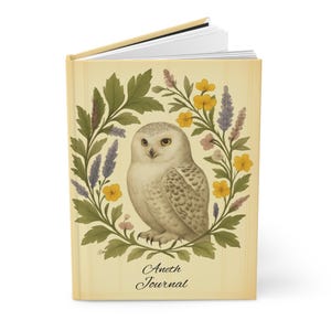 May include: A journal with a cream-colored cover featuring a detailed illustration of a snowy owl surrounded by a floral wreath. The wreath includes green leaves, yellow flowers, and purple accents. The text "Aneth Journal" is printed at the bottom.