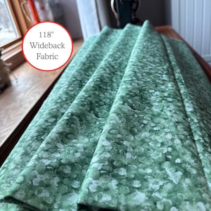 May include: Green and white patterned wideback fabric, 118 inches wide. The fabric is folded, showing the pattern of white spots on a green background. The fabric is on a wooden surface near a window.