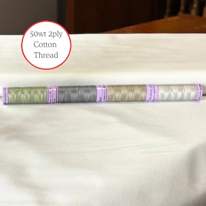 May include: Four spools of 50wt 2ply cotton thread arranged in a horizontal row. The spools feature shades of light green, grey, beige, and off-white, each wrapped with a purple label containing printed text details along the center of the spool.