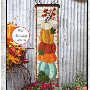 Könnte beinhalten: Ein vertikales Wandbehangmuster mit einem Stapel Kürbisse in Weiß, Gelb, Orange und Türkis. Das Design umfasst Herbstlaub und den Text "Wall Hanging Pattern" in einem roten Kreis. Der Hintergrund ist verwittertes, gewelltes Metall.