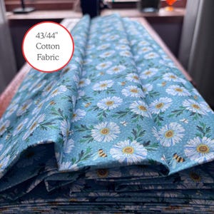 May include: A folded stack of 43/44" cotton fabric. The fabric features a light blue background with a repeating pattern of white daisies, green leaves, and small yellow bees. The text "43/44" Cotton Fabric" is in a red circle.