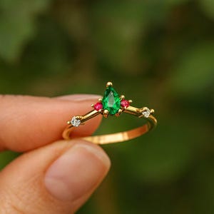 May include: A gold ring with a central pear-cut emerald, two small red gems, and two white diamonds. The ring's delicate band curves upwards. The ring is held against a blurred green background.