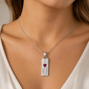 Può includere: Collana con pendente in argento con due targhette rettangolari, ciascuna incisa con un nome. Una gemma rossa a forma di cuore è centrata tra le targhette. La collana è indossata intorno al collo.