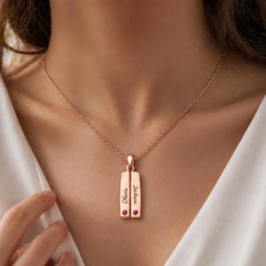 Può includere: Collana con pendente in oro rosa con due ciondoli rettangolari. Ogni ciondolo ha una piccola gemma rossa e un nome inciso. I nomi "Olivia" e "Jackson" sono visibili. La collana è indossata su una camicia bianca.