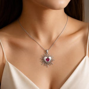 Puede incluir: Un collar de tono plateado con un colgante en forma de corazón y una piedra preciosa roja en el centro. El colgante está rodeado por un diseño de rayos con puntas metálicas y pequeñas esferas en los extremos, suspendido de una cadena fina.