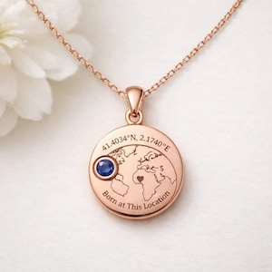 Puede incluir: Collar con colgante de oro rosa con un dije redondo con un diseño de mapa del mundo, una piedra preciosa azul y el texto "Born at This Location". El collar incluye coordenadas y un símbolo de corazón. La cadena es delicada.