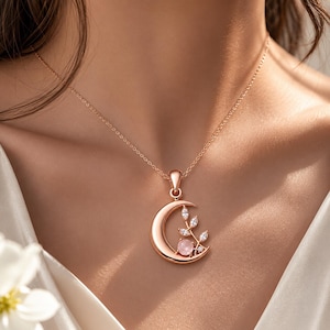 May include: Rose gold crescent moon pendant necklace with a pink gemstone and leaf accents. The necklace features a delicate chain and is a stylish piece of jewellery.