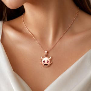 May include: A rose gold necklace featuring a pendant shaped like a smiling devil face with small horns. The pendant is accented with two small, round, vibrant pink gemstones set as the eyes, suspended from a delicate rose gold cable chain.