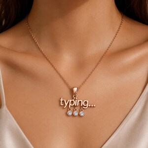 Puede incluir: Un collar de tono oro rosa con un colgante que muestra la palabra "typing..." en letras minúsculas. Tres pequeñas piedras redondas, transparentes y facetadas cuelgan de la parte inferior de las letras, cada una en un marco circular de oro rosa.