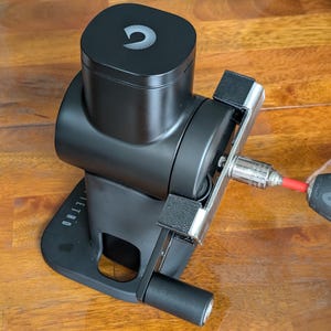 Op de afbeelding: Een zwart apparaat met een zilveren en rode component, mogelijk een garenwinder, wordt getoond op een houten oppervlak. Een draadloze boormachine is aan het apparaat bevestigd. Het apparaat heeft een vierkante bovenkant met een gebogen ontwerp.