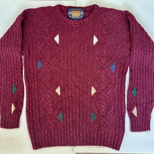 May include: A burgundy knitted jumper with a crew neck and long sleeves. The jumper features a diamond pattern with white, blue, and green geometric accents. The cuffs and hem have a white trim. The label at the neck reads "M.G. Sportswear".