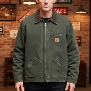 May include: Olive green work jacket with a brown corduroy collar. The jacket has a zip-up front, two front pockets, and a small patch on the chest. The jacket is a casual outerwear piece.