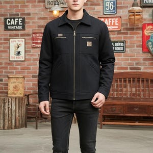 May include: A black jacket with a zipper closure, two chest pockets, and a collared neckline. The jacket has small patches on the chest and upper arm. The person is wearing black jeans. The background is a brick wall with vintage signs.