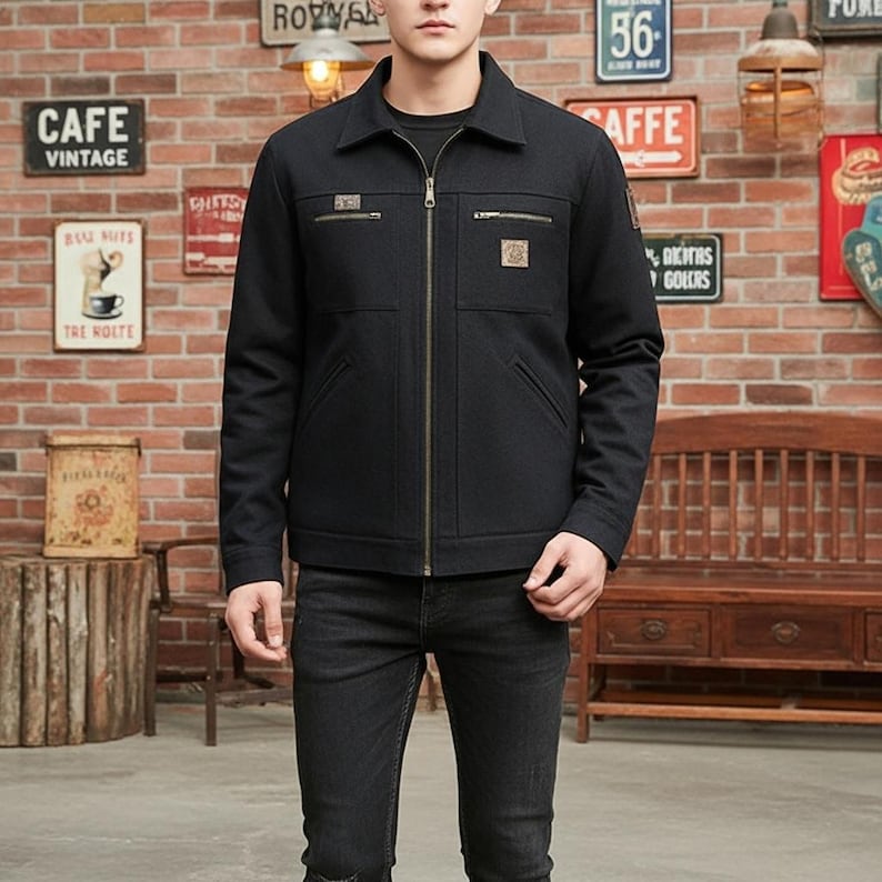 May include: A black jacket with a zipper closure, two chest pockets, and a collared neckline. The jacket has small patches on the chest and upper arm. The person is wearing black jeans. The background is a brick wall with vintage signs.
