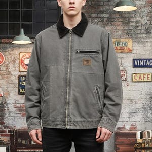 May include: A gray jacket with a black collar and a zipper closure. The jacket has a small pocket on the chest and a patch with text. The jacket is worn over a black shirt.