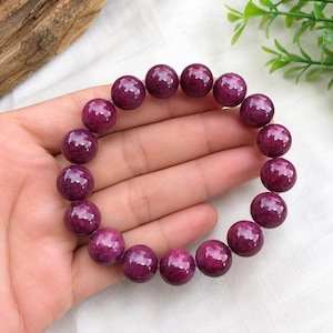 Puede incluir: Una pulsera circular compuesta por cuentas esféricas lisas y pulidas de color magenta intenso. Las cuentas son de tamaño uniforme y están ensartadas en un cordón elástico, creando un bucle continuo de esferas de piedra preciosa rosa oscuro.