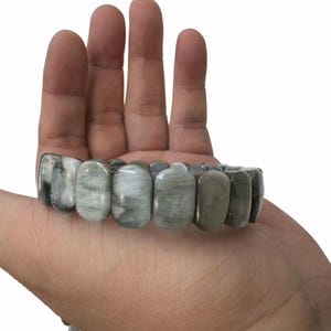 May include: A stretch bracelet composed of rectangular, polished stones in shades of gray, white, and green. The bracelet is displayed on a hand, showcasing the natural variations in the stone's color and texture. The stones are approximately 1 inch in length.