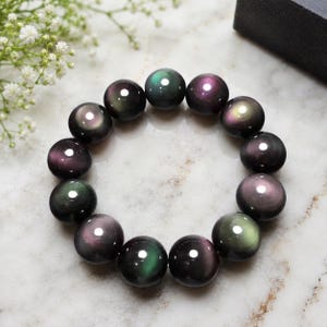 Op de afbeelding: Een cirkelvormige armband bestaande uit gladde, gepolijste, bolvormige kralen van donker obsidiaan. Elke kraal vertoont een uitgesproken iriserende glans met verschuivende accenten van groen, paars en goud, die het licht op hun oppervlak reflecteren.