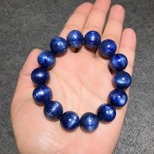 Puede incluir: Una pulsera de cuentas compuesta por piedras preciosas de cianita redondas y pulidas. Las cuentas exhiben un color azul profundo y vibrante con vetas chatoyantes visibles y un acabado brillante y reflectante en forma circular.