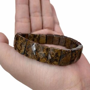 Könnte beinhalten: Ein braunes und beigefarbenes Edelsteinarmband mit rechteckigen, facettierten Perlen. Das Armband wird in einer Hand vor weißem Hintergrund gehalten. Die Perlen haben ein gesprenkeltes Aussehen mit Farb- und Texturvariationen.