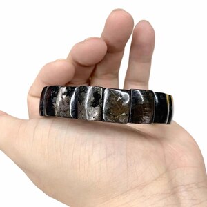 Puede incluir: Una pulsera negra y gris con piedras rectangulares pulidas. La pulsera se sostiene en una mano sobre un fondo blanco. Las piedras tienen un aspecto metálico reflectante, y la pulsera parece estar hecha de materiales naturales.